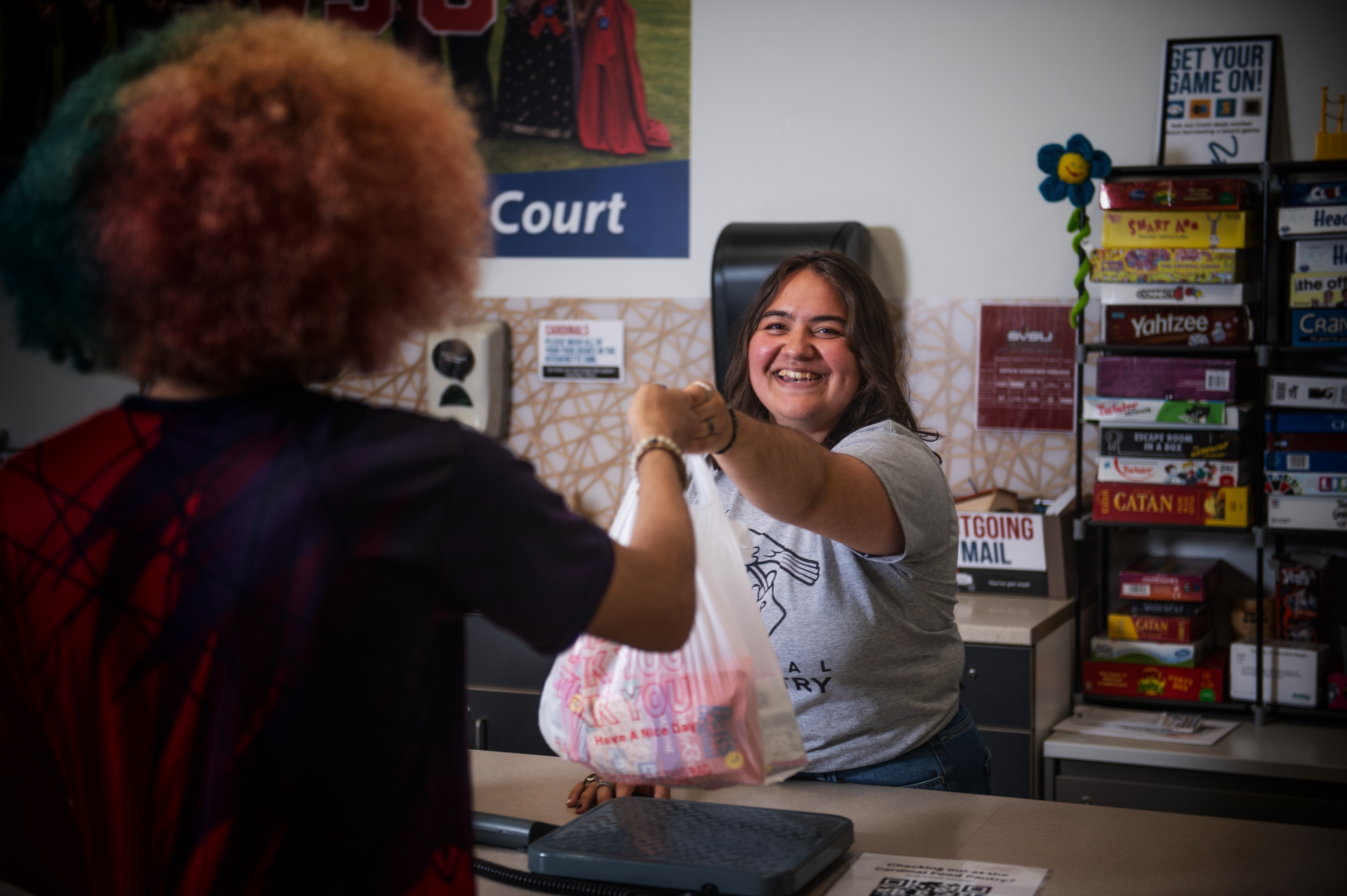 One student holds out a bag of groceries to hand it across the counter to another student receiving the bag.